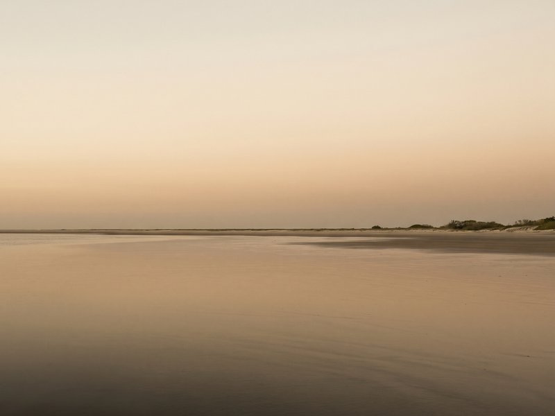 Horizonte amplo sobre praia, areia molhada espelhando o céu em tons quentes.