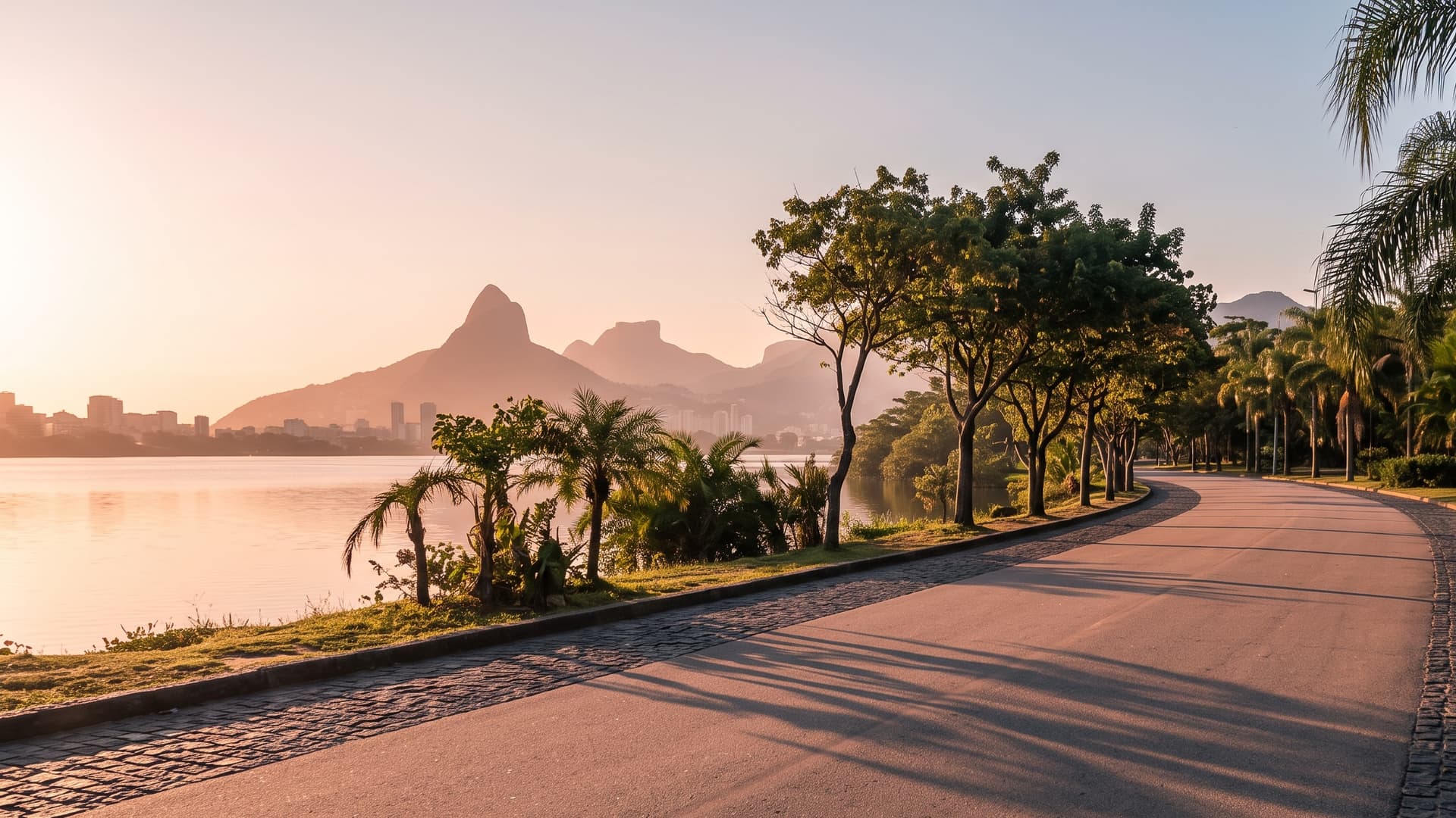 Via arborizada vazia com faixa de paralelepípedos, curvando junto a uma lagoa com morros ao fundo e sombras longas de árvores sobre o asfalto